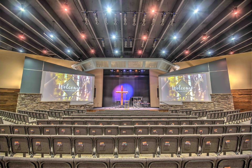 Picture of Pulpit Rock Auditorium Renovation, pendant lights, large worship seating, balcony viewing area in churches, glass partition in church assembly, exposed rafters in church interior, modern church sanctuary, wood trim in interior church building. Bright church auditorium. Large TV in church auditorium. Center stage church auditorium.